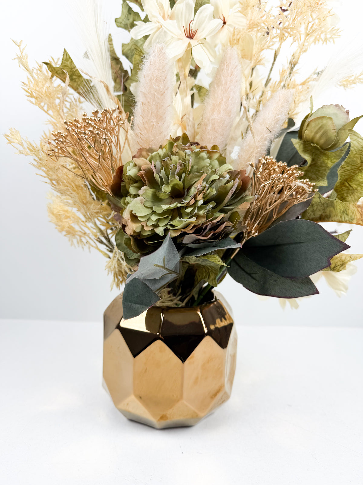 Arrangement de fleurs séchées vert doux dans un vase géométrique doré facetté.