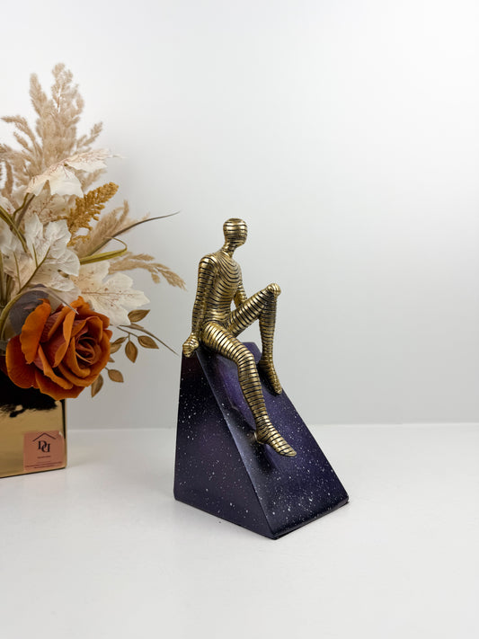 Sculpture décorative contemporaine d'un personnage doré texturé assis sur un socle géométrique bleu nuit effet ciel étoilé.