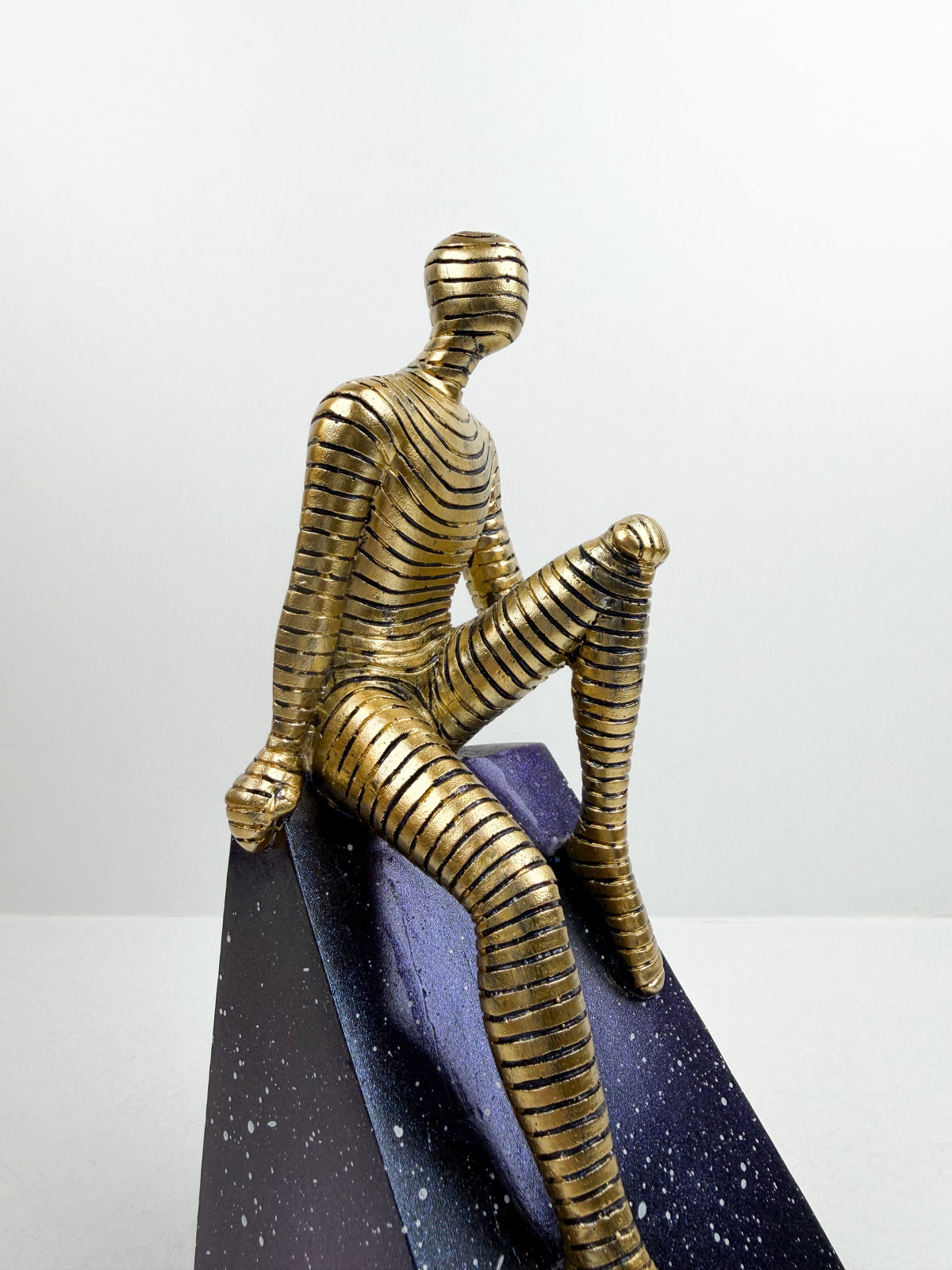 Sculpture décorative contemporaine d'un personnage doré texturé assis sur un socle géométrique bleu nuit effet ciel étoilé.
