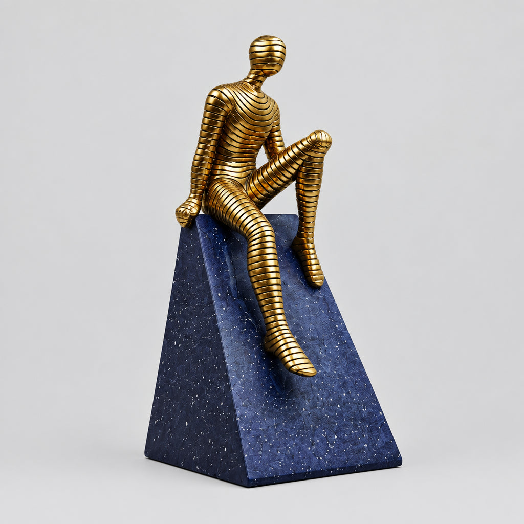 Sculpture décorative contemporaine d'un personnage doré texturé assis sur un socle géométrique bleu nuit effet ciel étoilé.